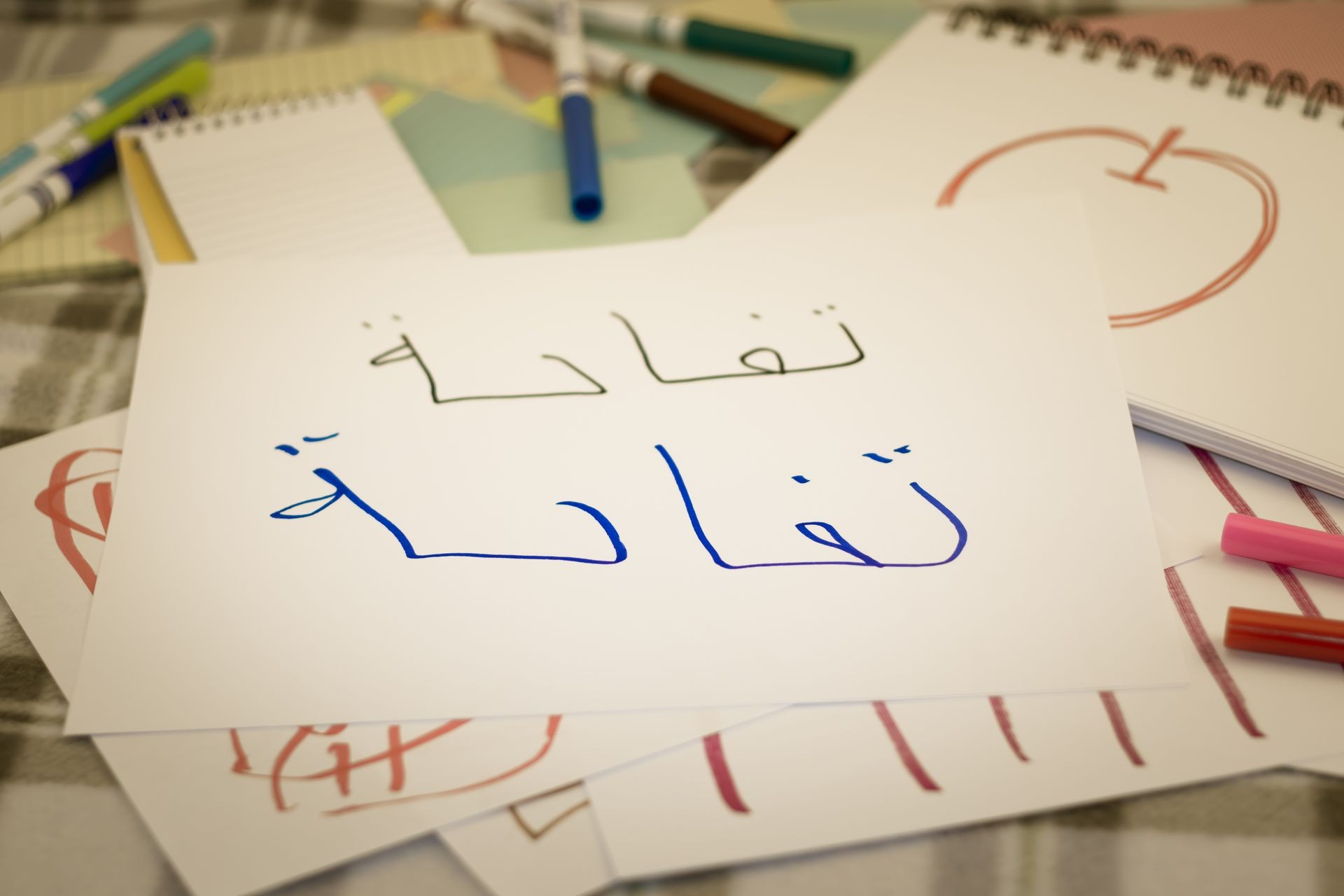 Paper with Arabic handwriting and markers scattered on a table, alongside sketchpads with drawings.